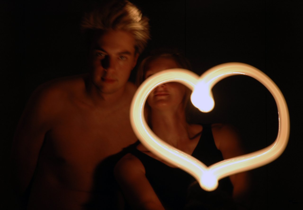Two people stand in a dark setting while one draws a glowing heart shape using light.