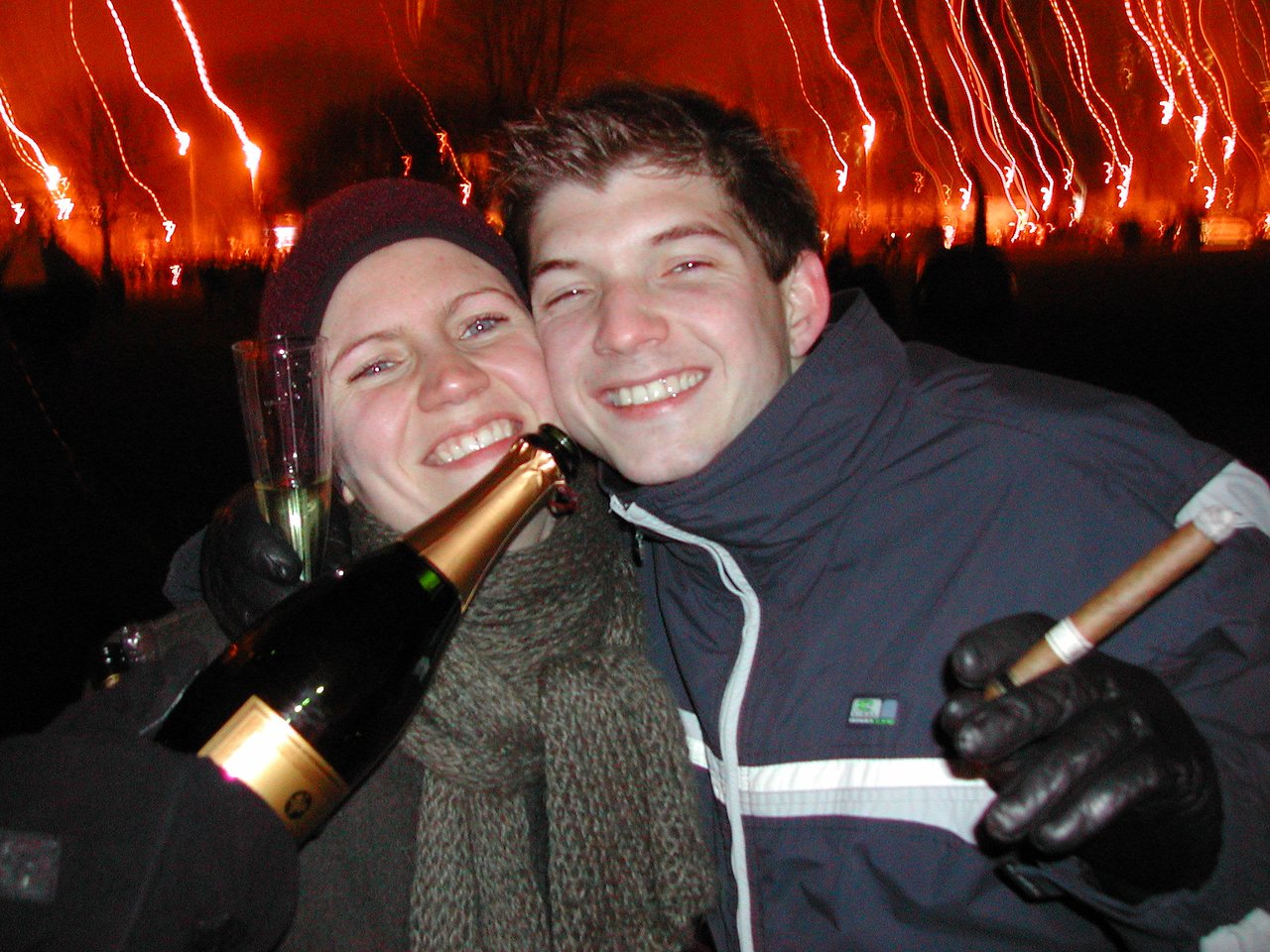 Two people smiling and celebrating New Year's Eve, holding champagne and a cigar in a festive outdoor setting.