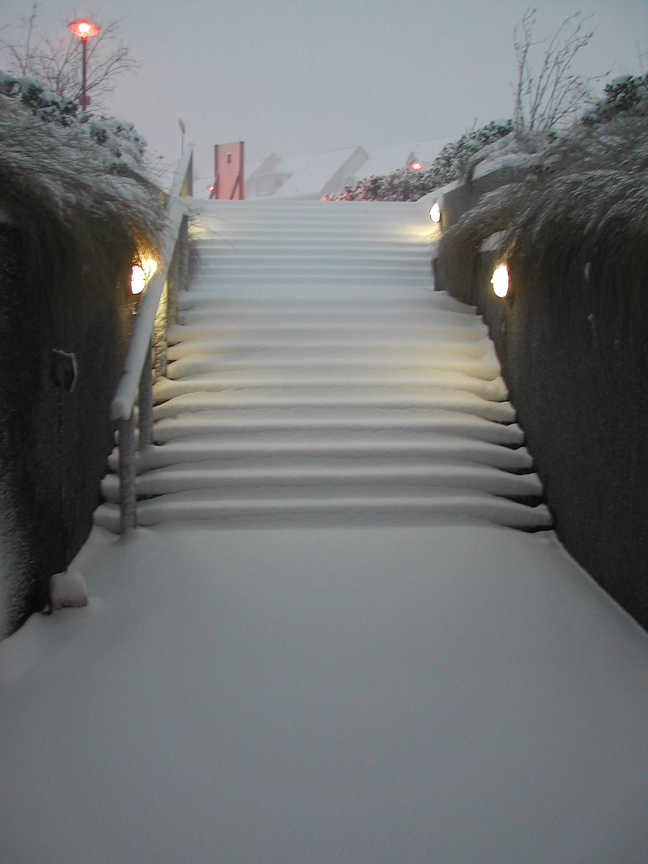 A snow-covered outdoor staircase with handrails and lights on both sides, leading up to buildings in the background.