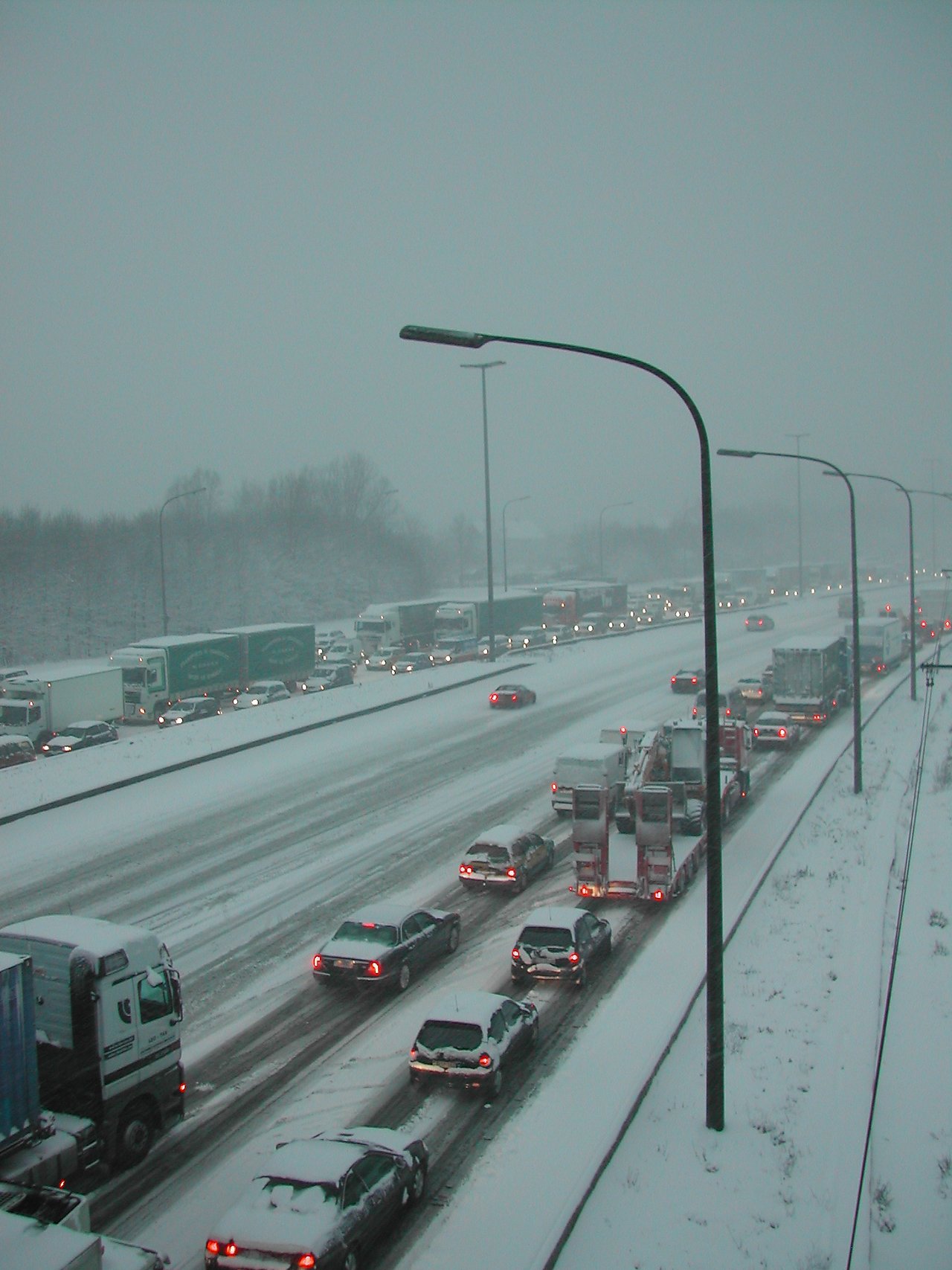 Snow-covered highway with heavy traffic, including cars and trucks, moving slowly in both directions during snowy weather.