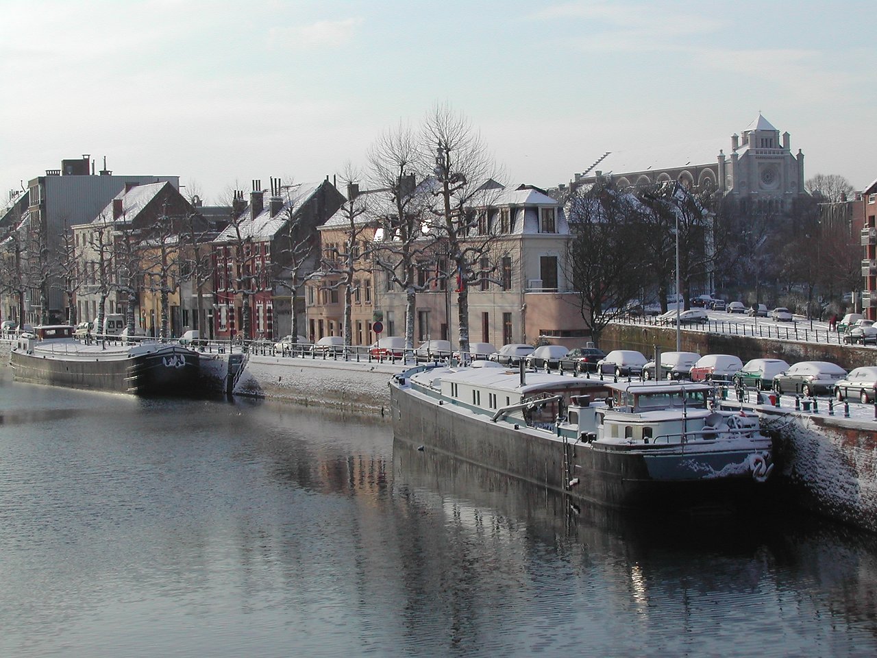 Snow-covered boats docked along a canal in Ghent, with colorful buildings and a church in the background.