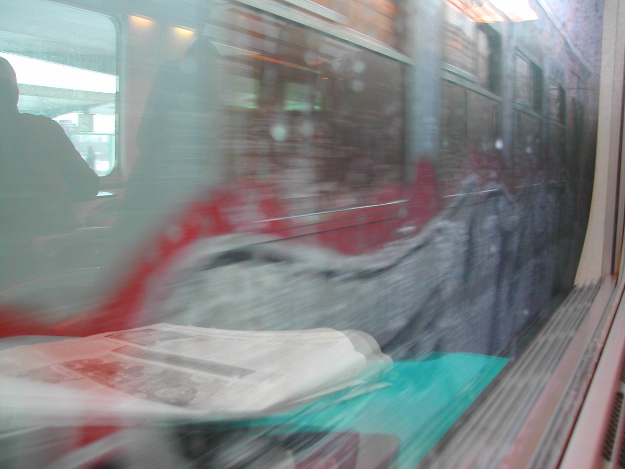 A train passes by the window, reflecting a newspaper and a passenger inside another train.