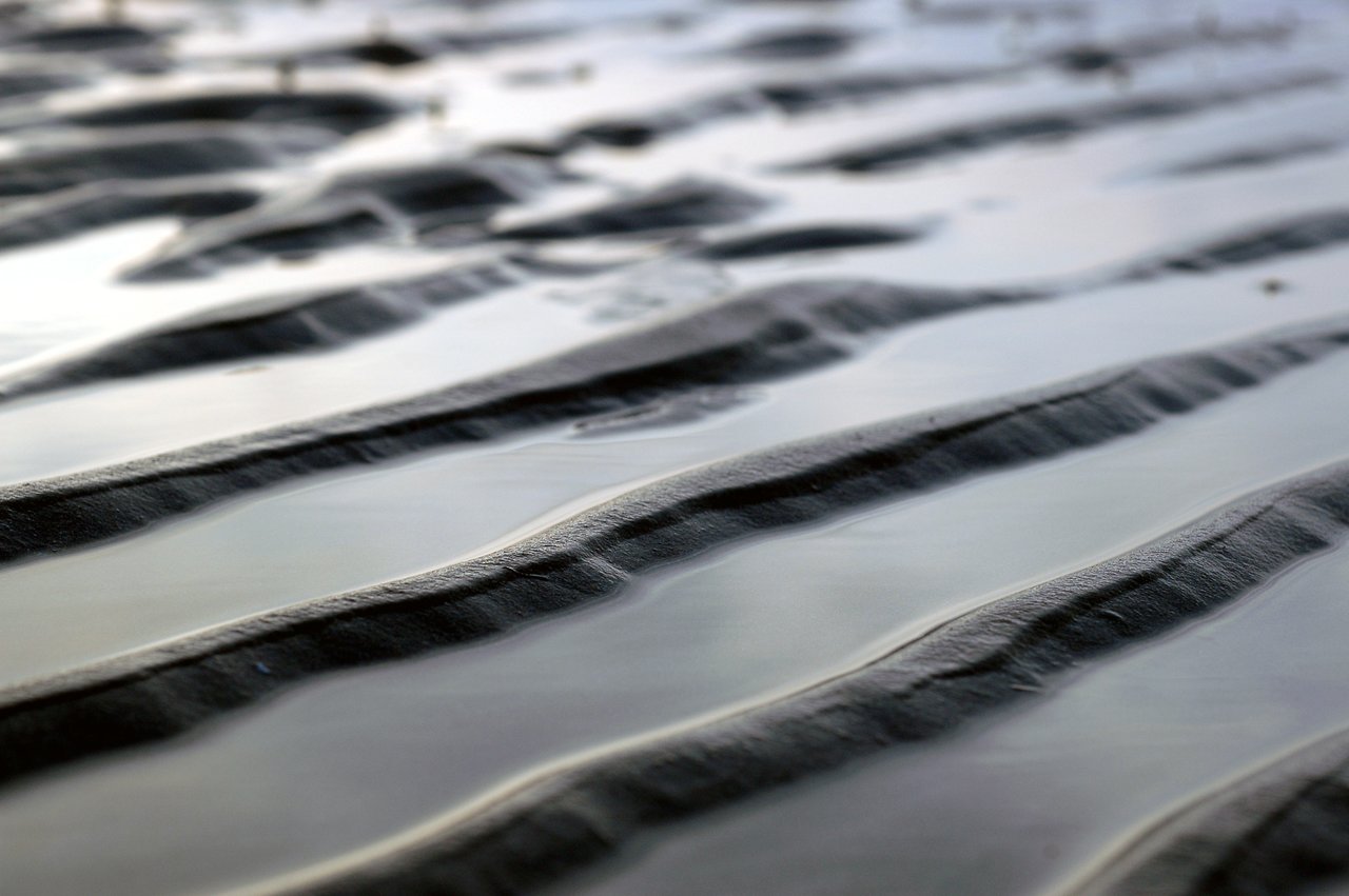 Close-up of wet sand with rippled patterns formed by the tide, partially covered by shallow water.