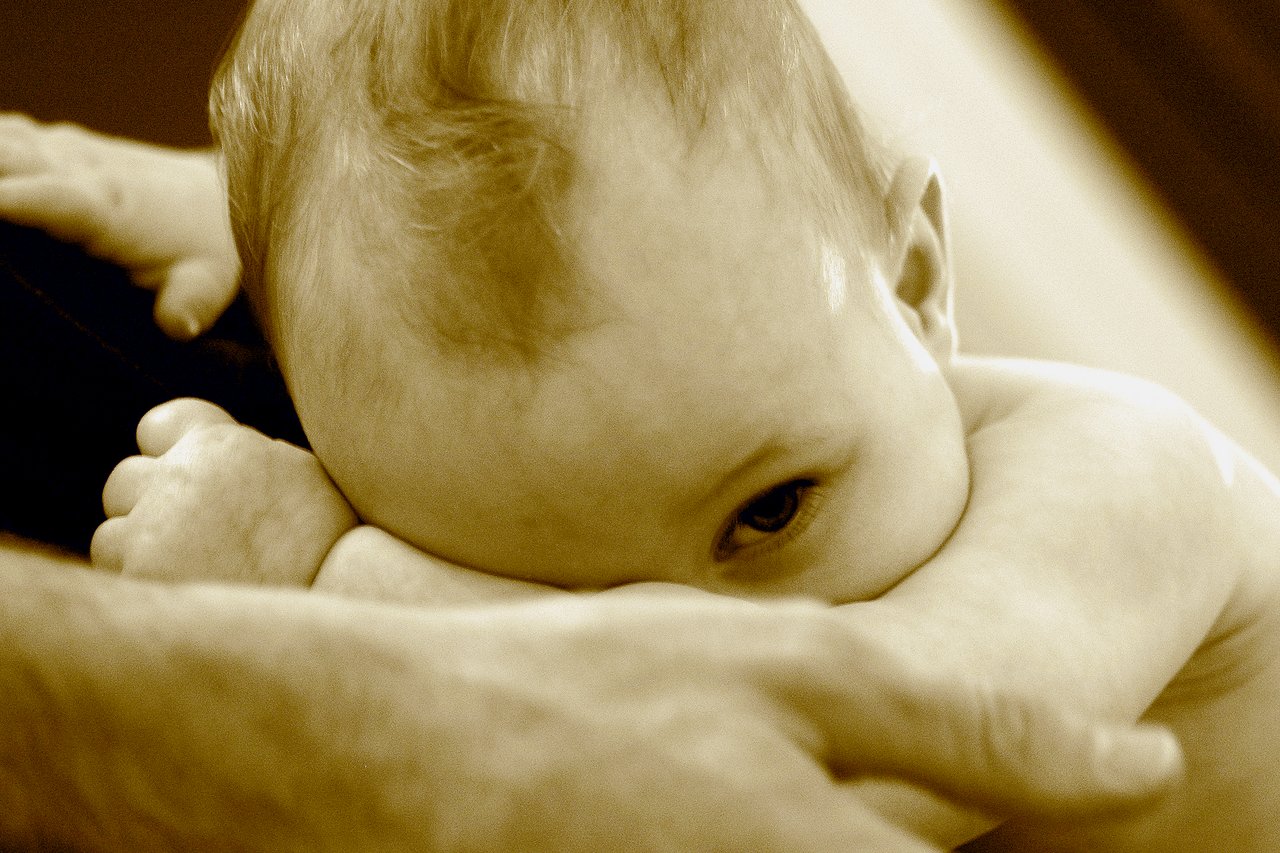 A baby rests their head on an adult's arm, gazing to the side with a calm expression.