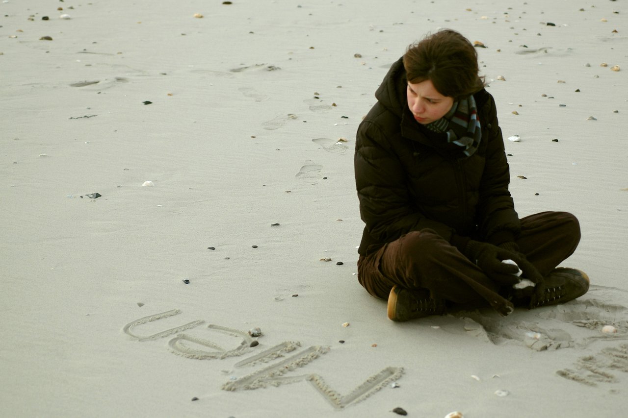 A person in a dark coat sits on a sandy beach, looking at the word "Mien" written in the sand.