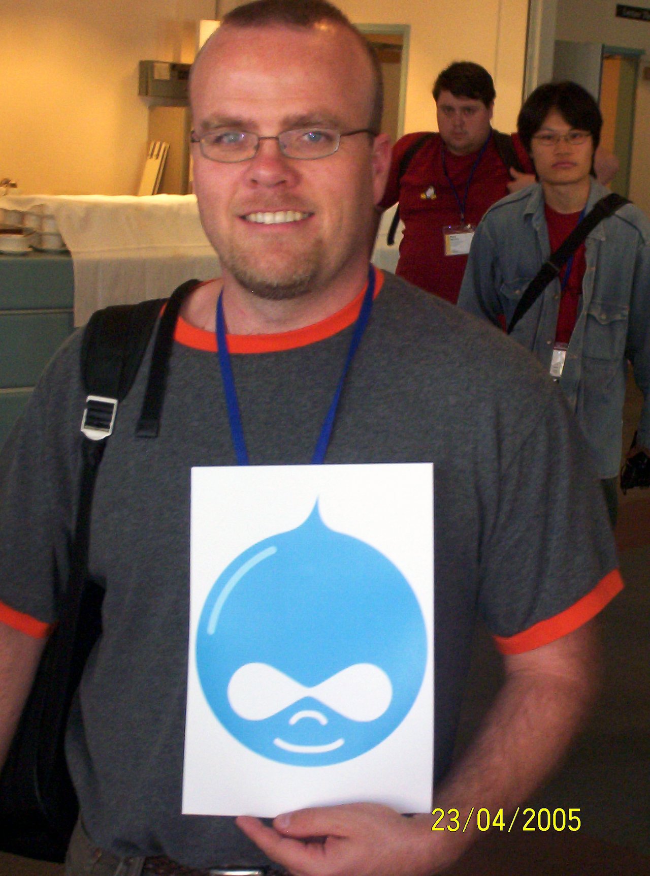 A man holds a sign with the Drupal logo at a Linux conference, smiling at the camera.