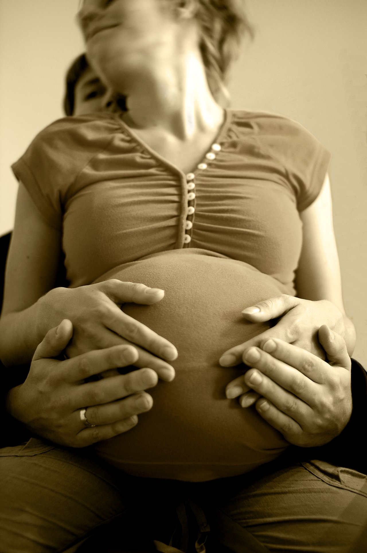 A pregnant woman sits while hands, likely hers and her partner's, gently cradle her belly.