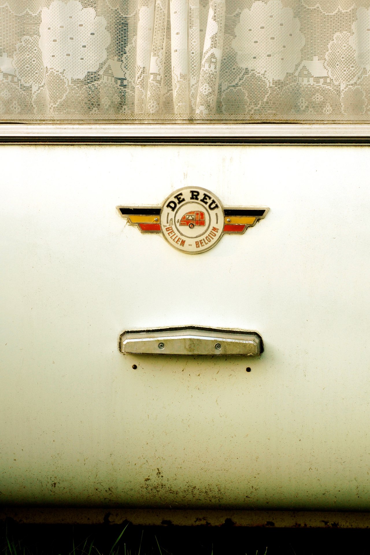 Close-up of a Belgian trailer door featuring a "De Reu" emblem, metal handle, and a lace-curtained window.