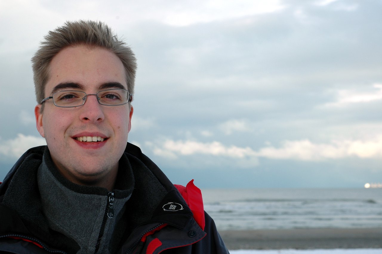 A man wearing glasses and a winter jacket smiles while standing on a beach with the ocean behind him.