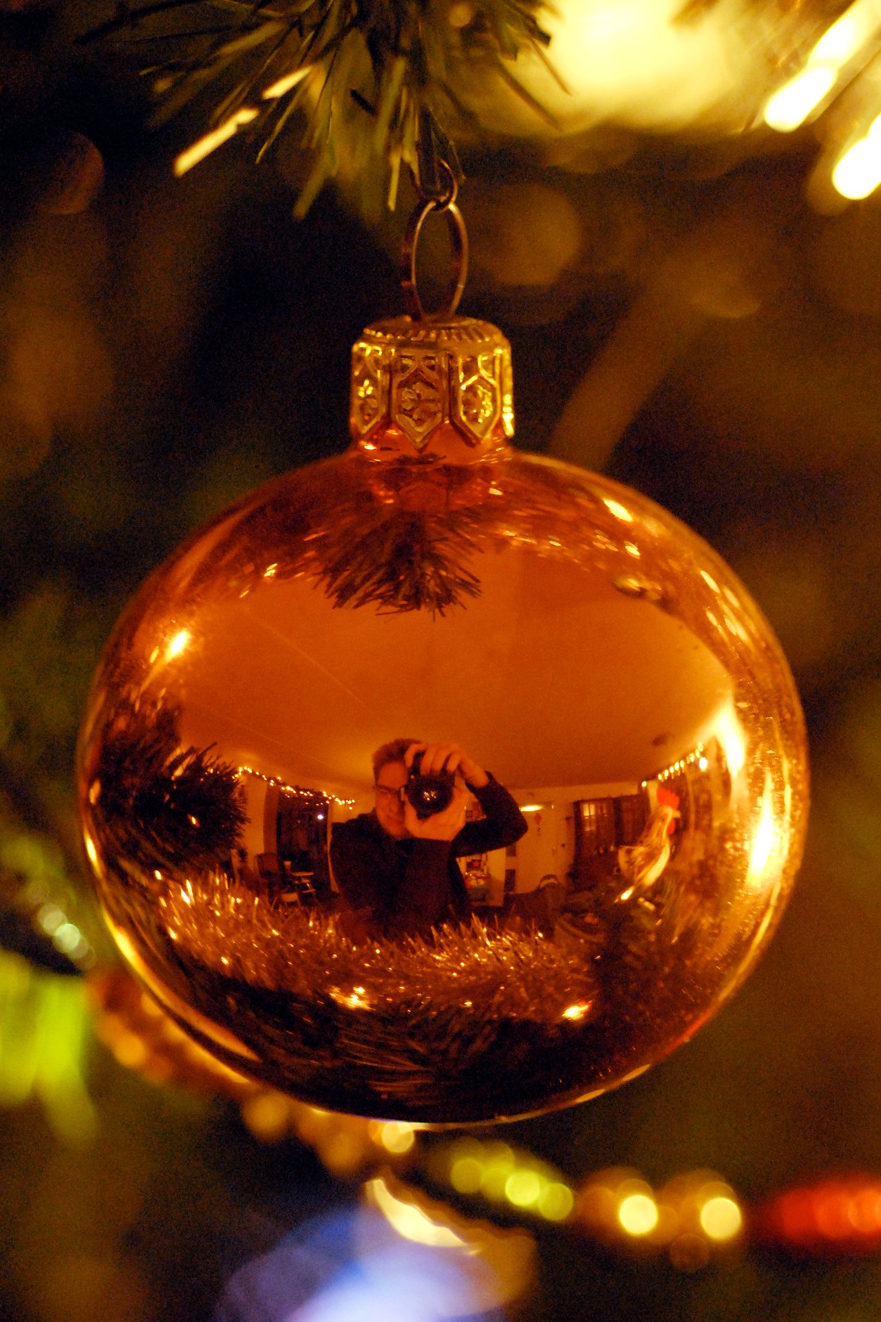 A person taking a photo is reflected in a shiny gold Christmas ornament hanging on a decorated tree.
