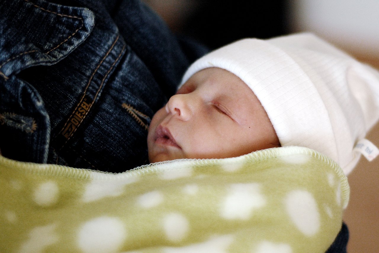 A newborn baby sleeps, wrapped in a soft green blanket and wearing a white hat.