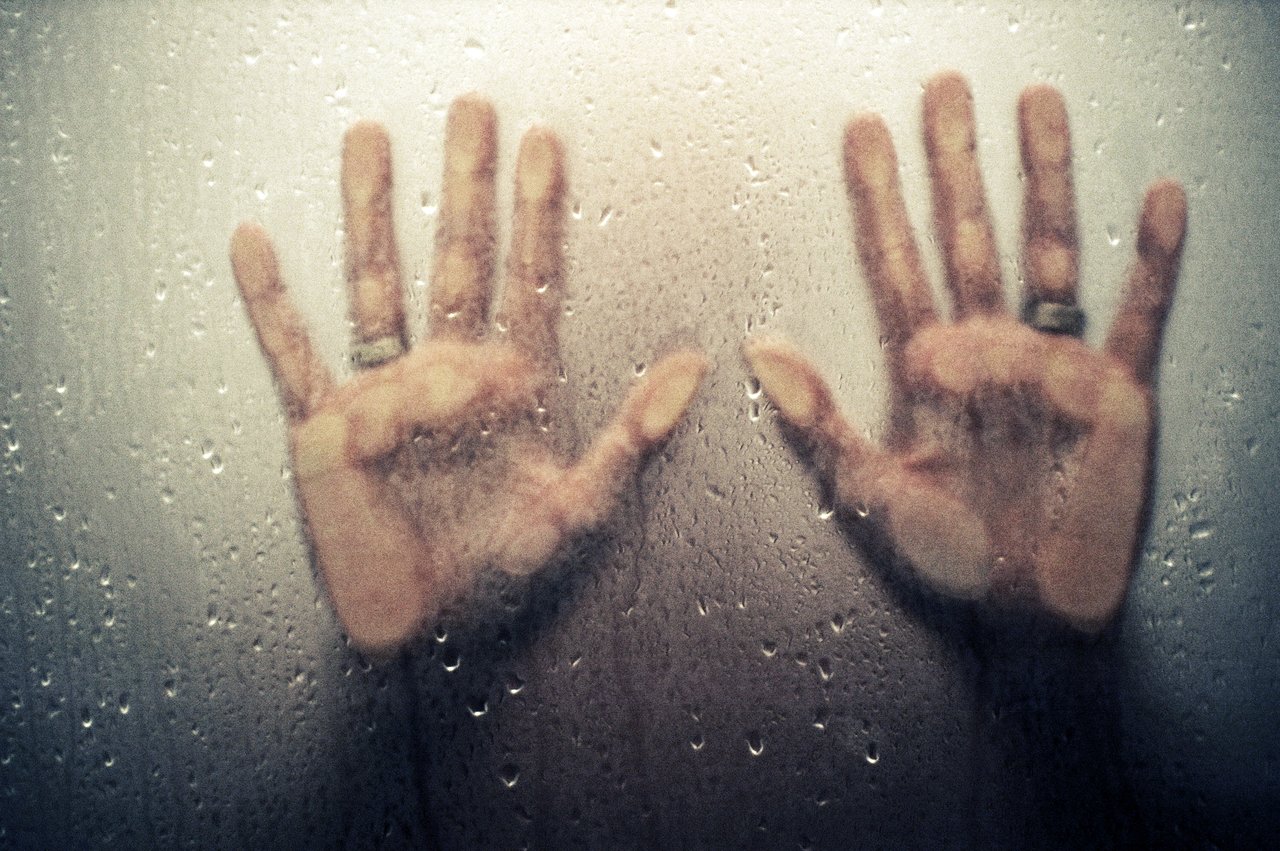 Two hands with fingers spread press against a wet, foggy glass surface, partially obscured by condensation and water droplets.