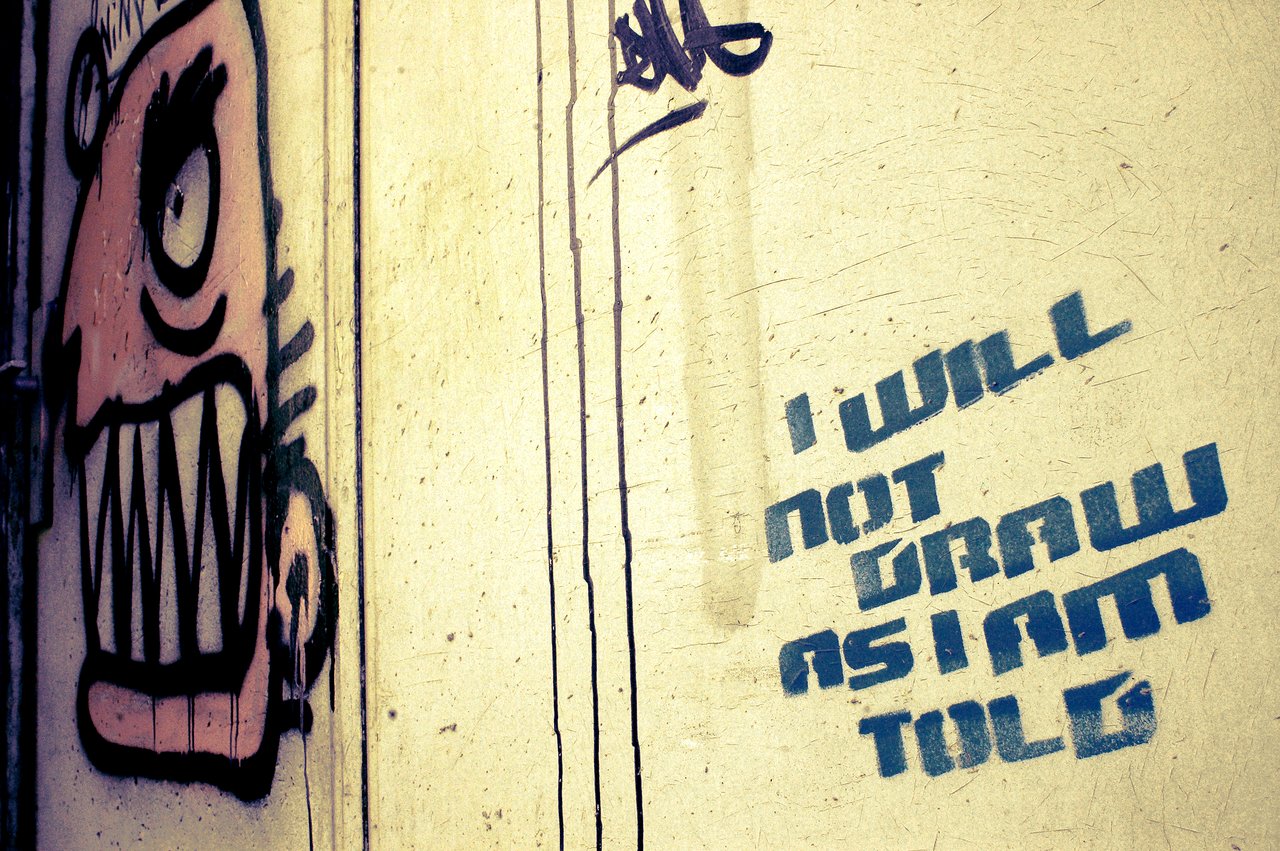 Graffiti of a cartoonish face with sharp teeth next to stenciled text reading, "I will not draw as I am told.