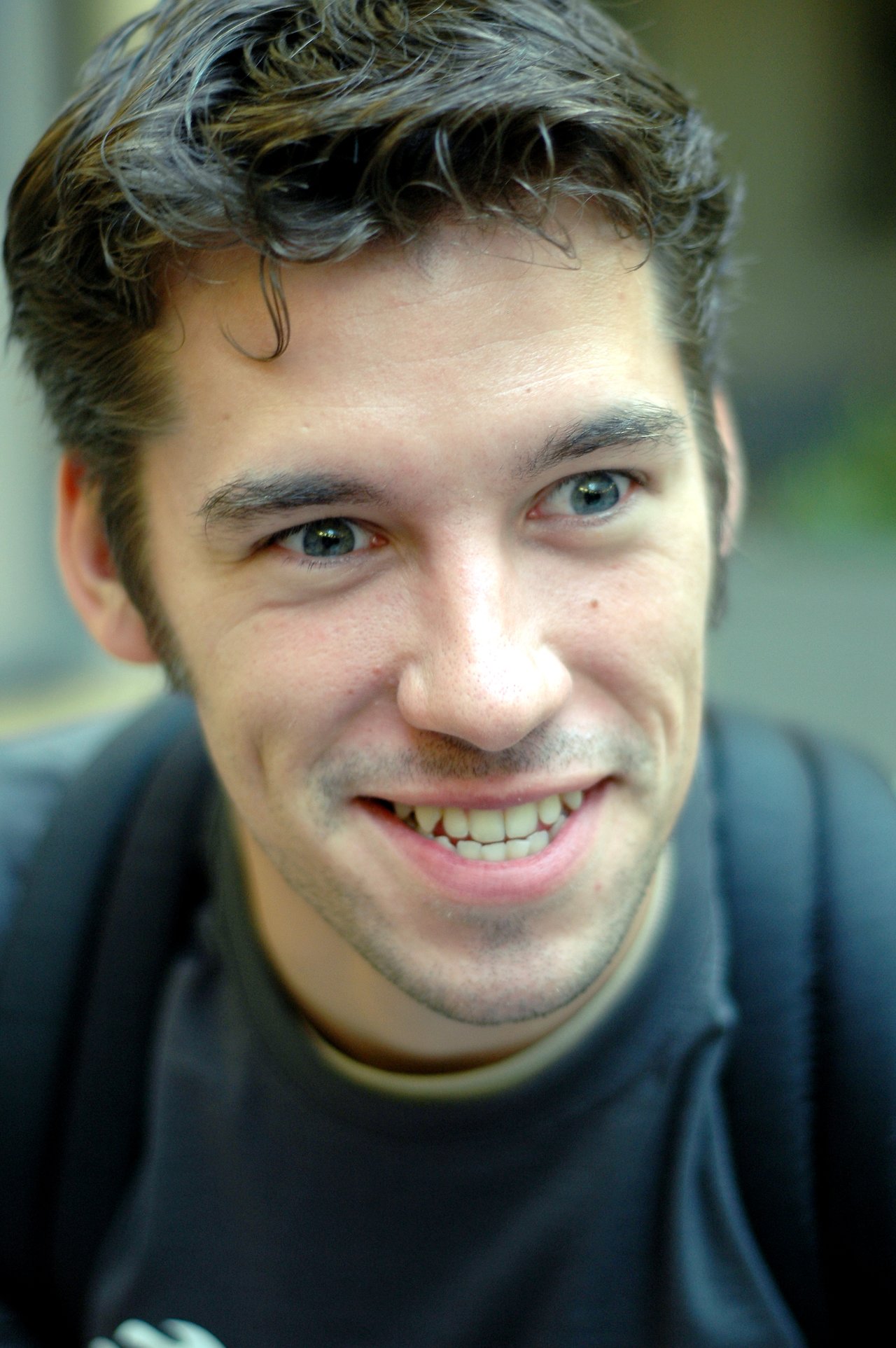 A young man with short dark hair and a backpack smiles while looking slightly off-camera.