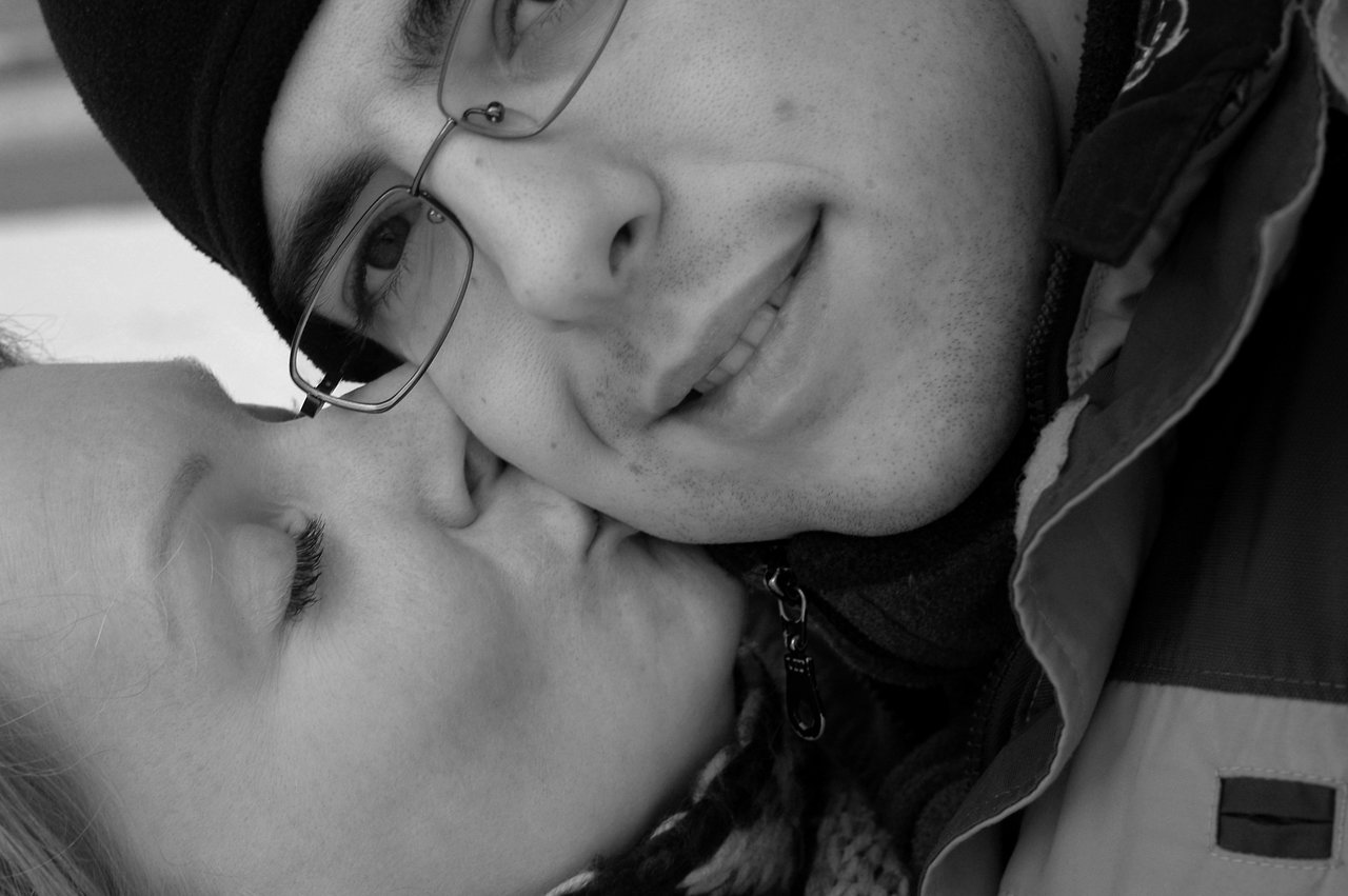 A woman kisses a smiling man on the cheek while he looks at the camera.