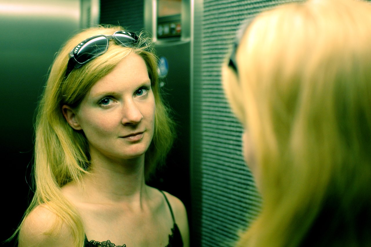A woman with sunglasses on her head looks at the camera in an elevator, with her reflection visible.