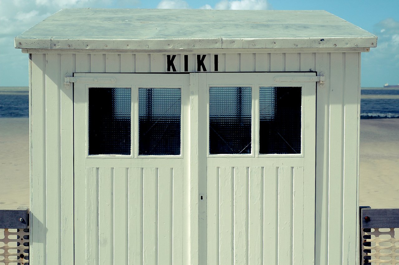 A small white beach hut named 'Kiki' with closed doors and windows, facing the sandy shore.