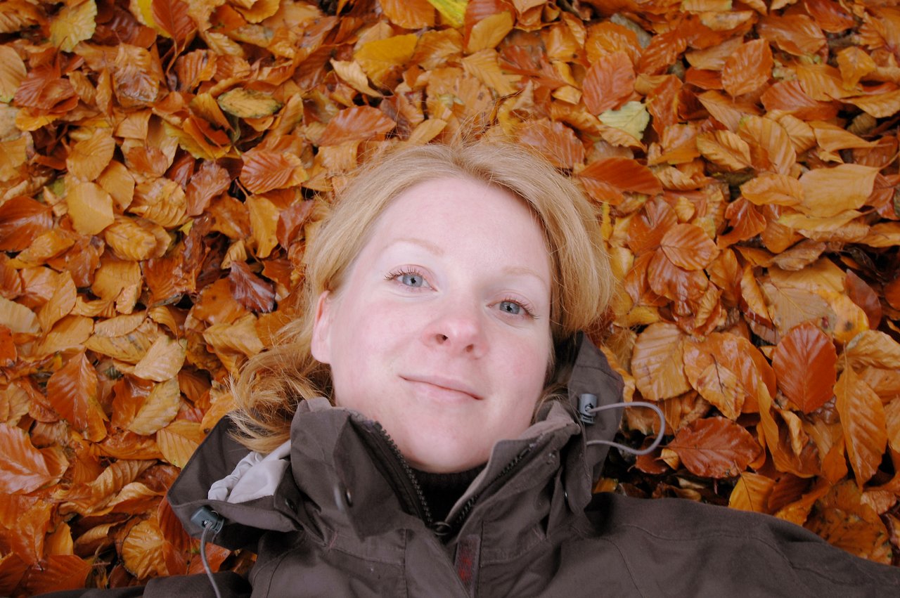 A woman with blonde hair lies on autumn leaves, wearing a dark jacket and looking directly at the camera.