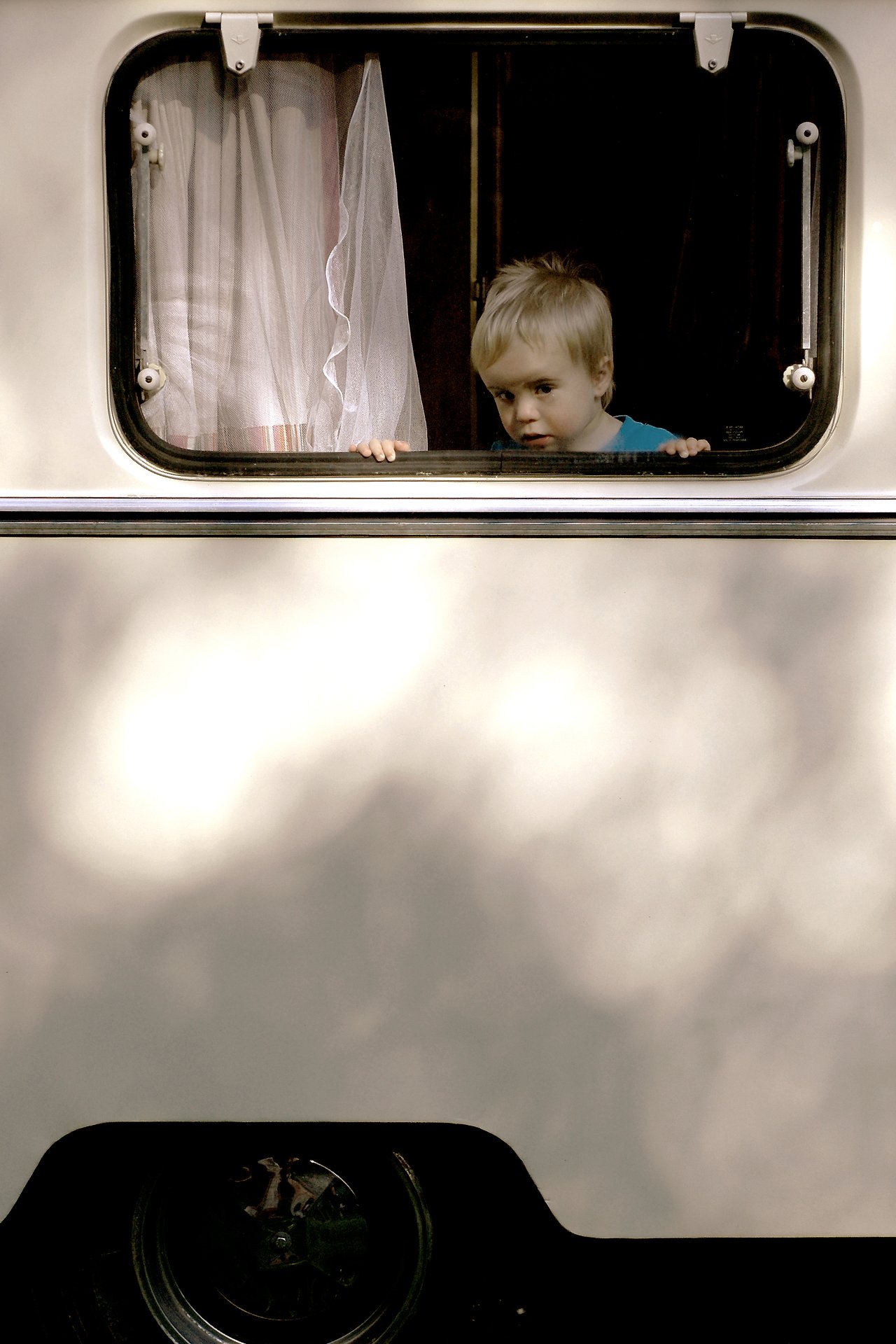 A young boy in a blue shirt looks out from a camper window, holding the frame with both hands.