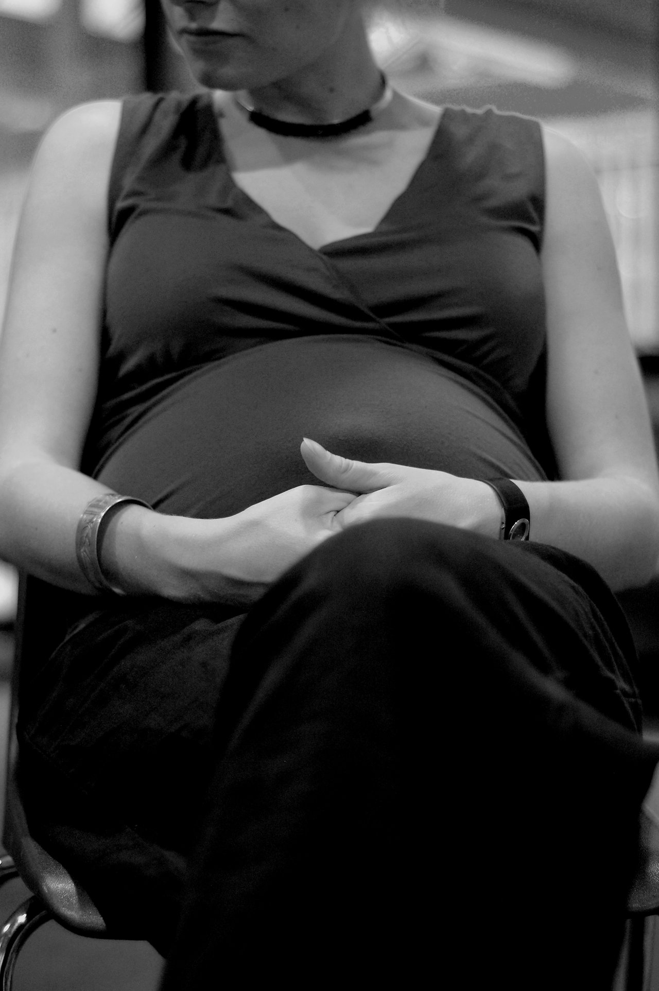 A pregnant woman sits with her hands on her belly, wearing a sleeveless dress, bracelets, and a watch.