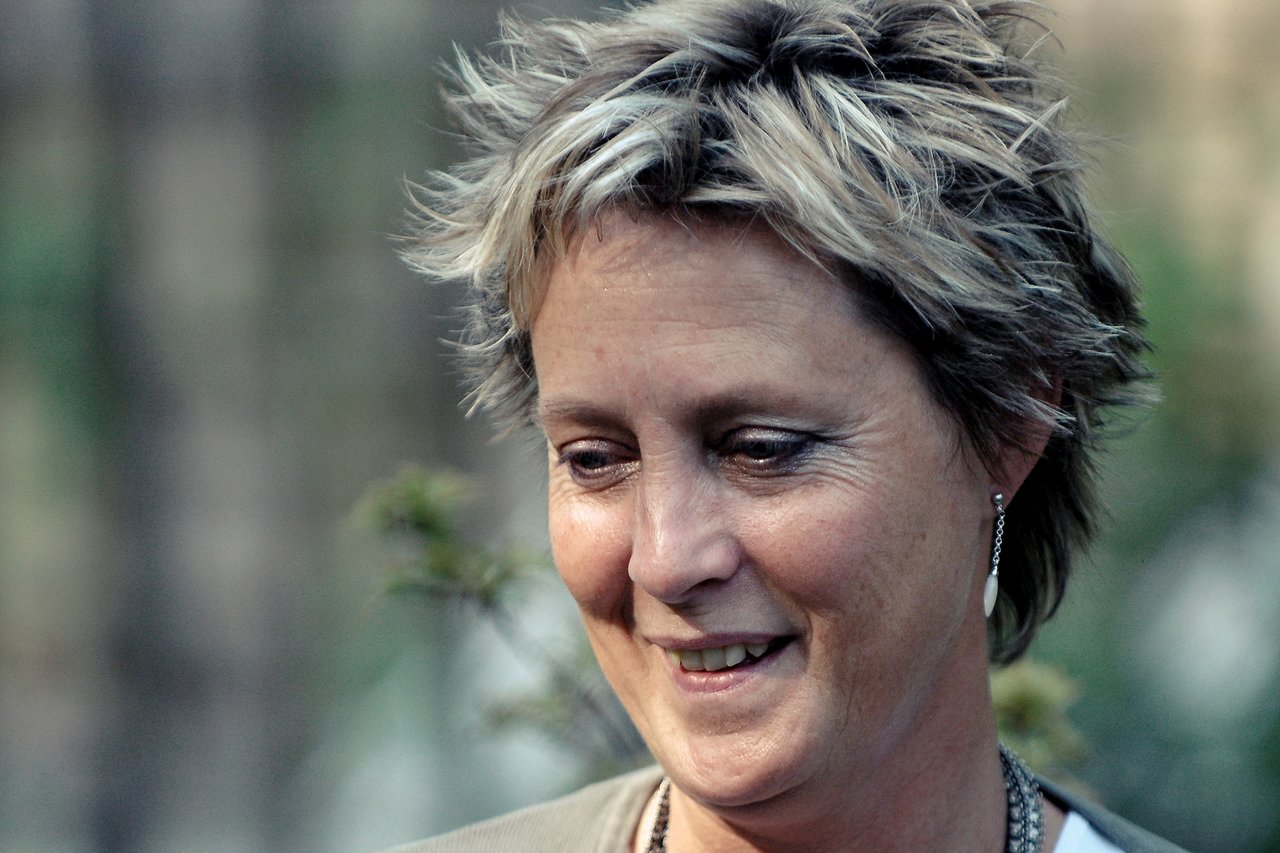 A woman with short blonde hair smiles gently while looking downward in an outdoor setting.