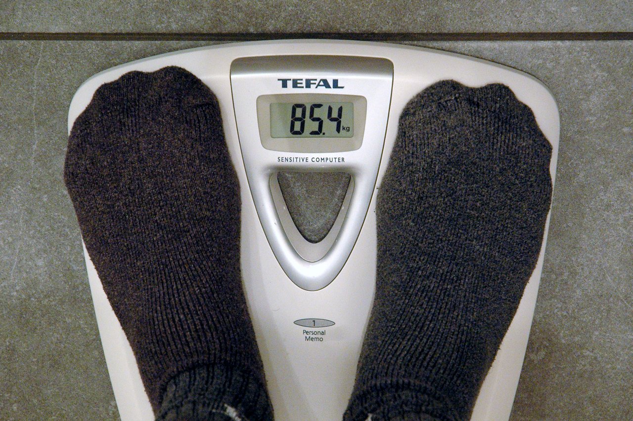 A person wearing dark socks stands on a digital scale showing a weight of 85.