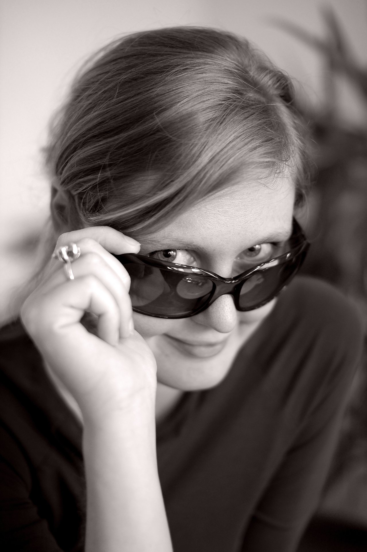 A woman lifts her sunglasses slightly and looks at the camera with a subtle smile.