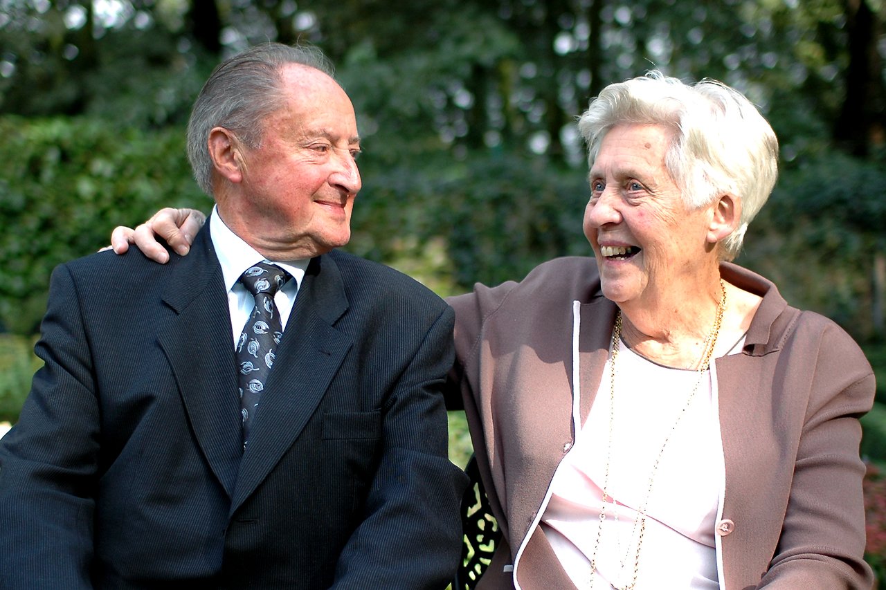An elderly couple sits together, smiling at each other while celebrating their 55th wedding anniversary outdoors.