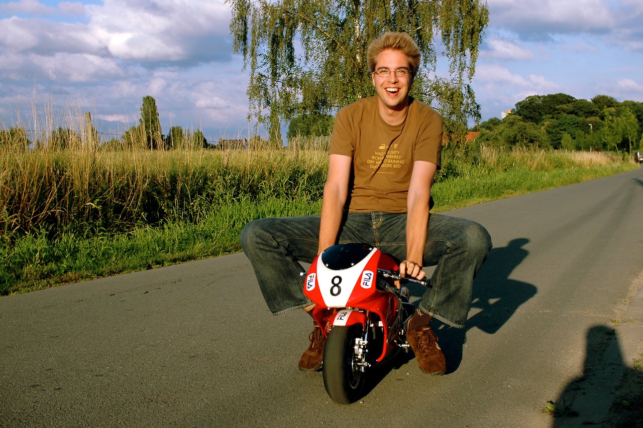 A man in a brown shirt smiles while sitting on a small red pocket bike on a rural road.