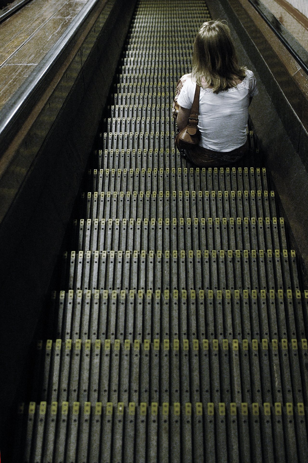 A person sits on an escalator step, facing away, wearing a white shirt and carrying a brown shoulder bag.