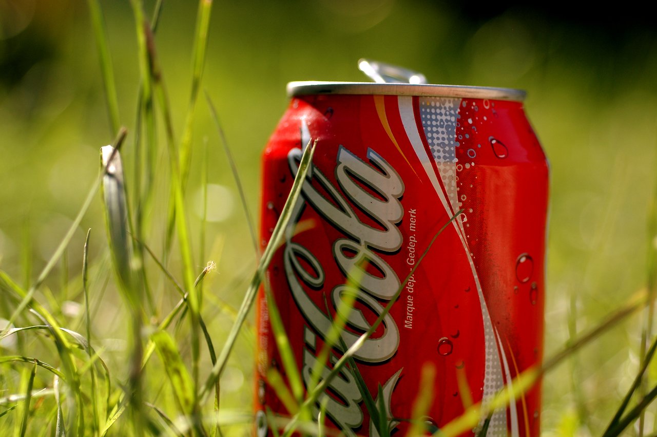An open Coca-Cola can sits on the grass, surrounded by green blades in bright sunlight.