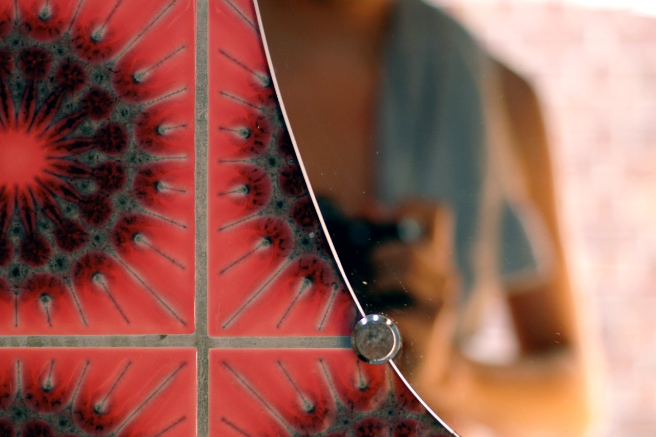 Close-up of red patterned tiles next to a mirror reflecting a person holding a camera.