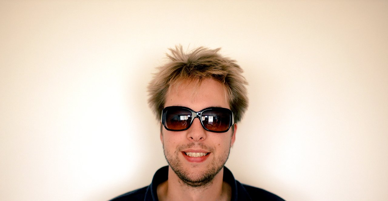 A man with messy hair, stubble, and sunglasses smiles at the camera against a plain background.
