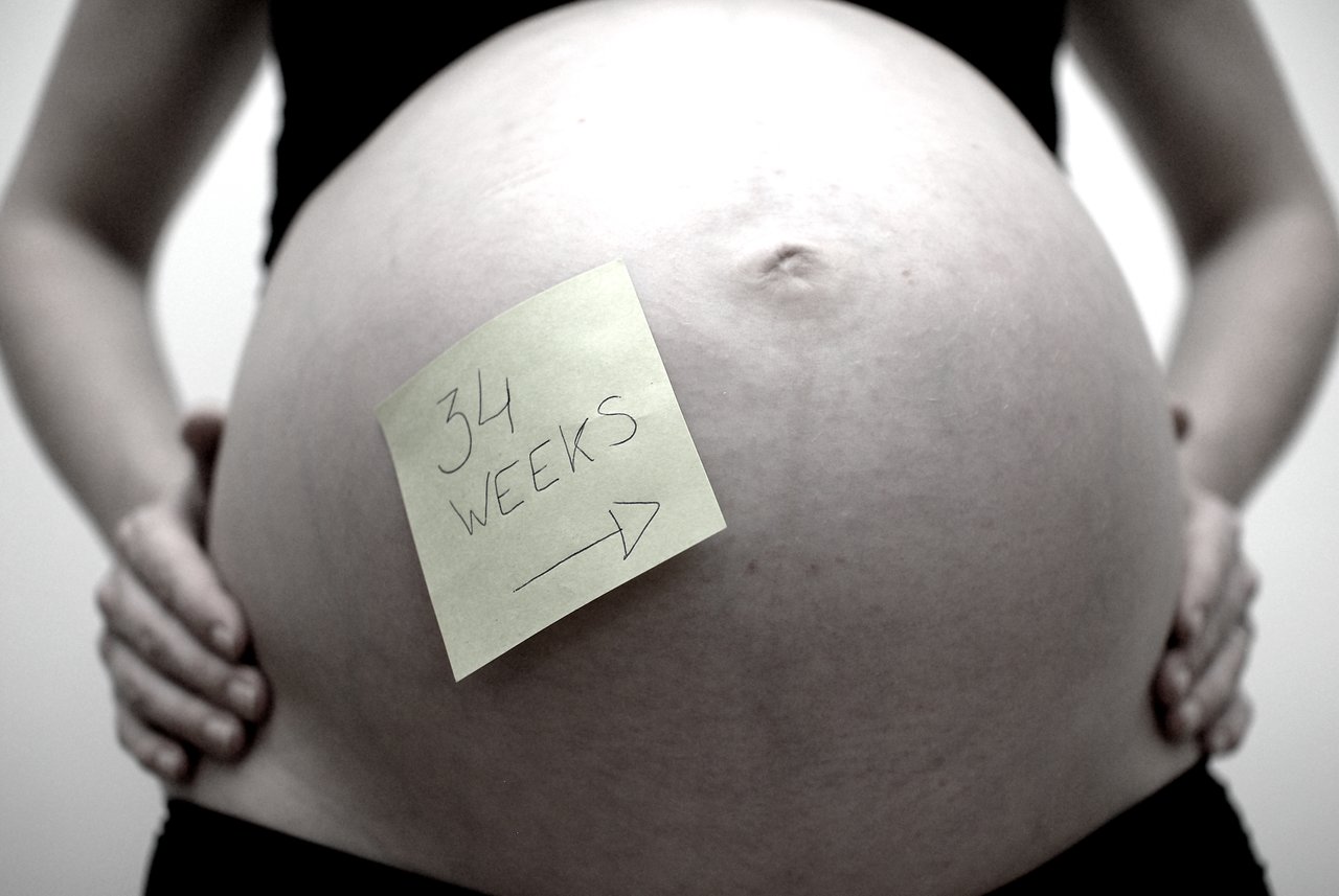 A pregnant person holds their belly with a sticky note that reads "34 weeks" attached to it.