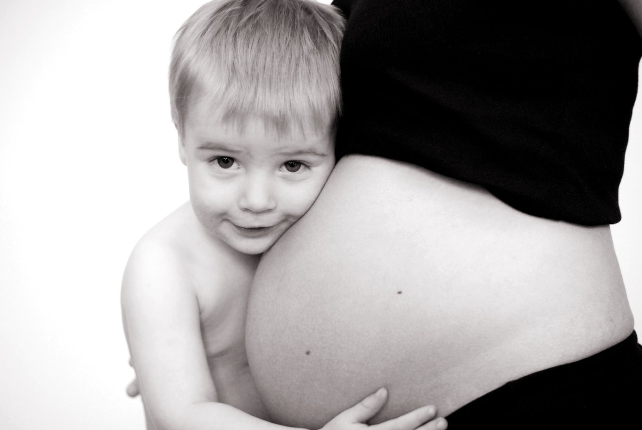 A young child hugs and rests his face against his pregnant mother's belly, looking at the camera.