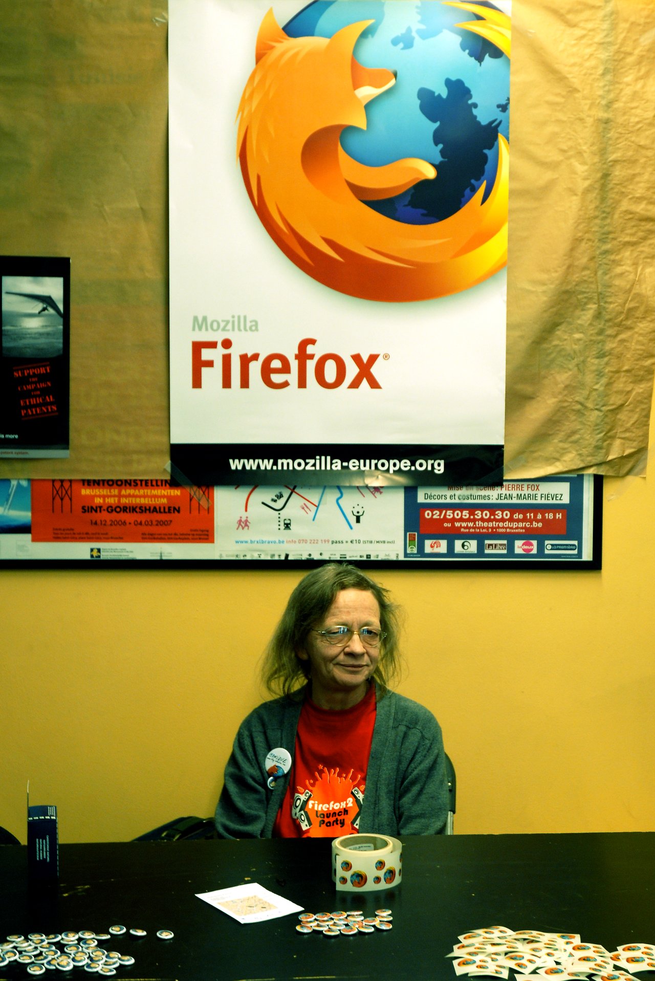 A woman in a Firefox T-shirt sits at a booth with stickers and buttons, promoting Mozilla at FOSDEM.