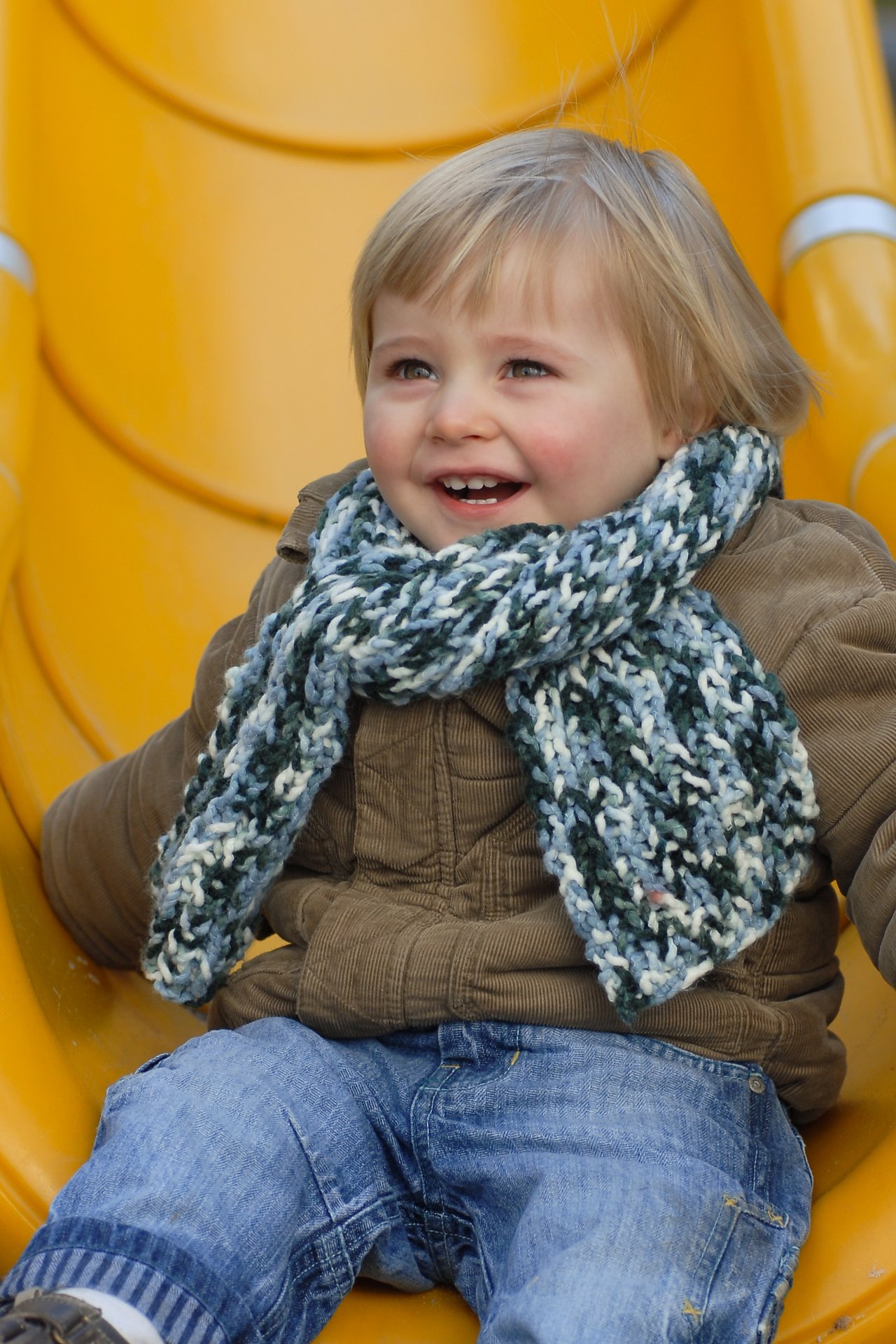 A smiling young child with a scarf and jacket sits on a yellow slide, looking happy.