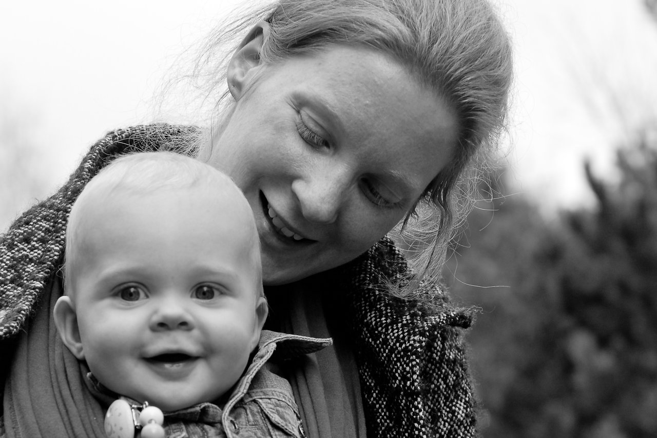 A smiling baby in the foreground with a woman behind them, looking down and smiling warmly.