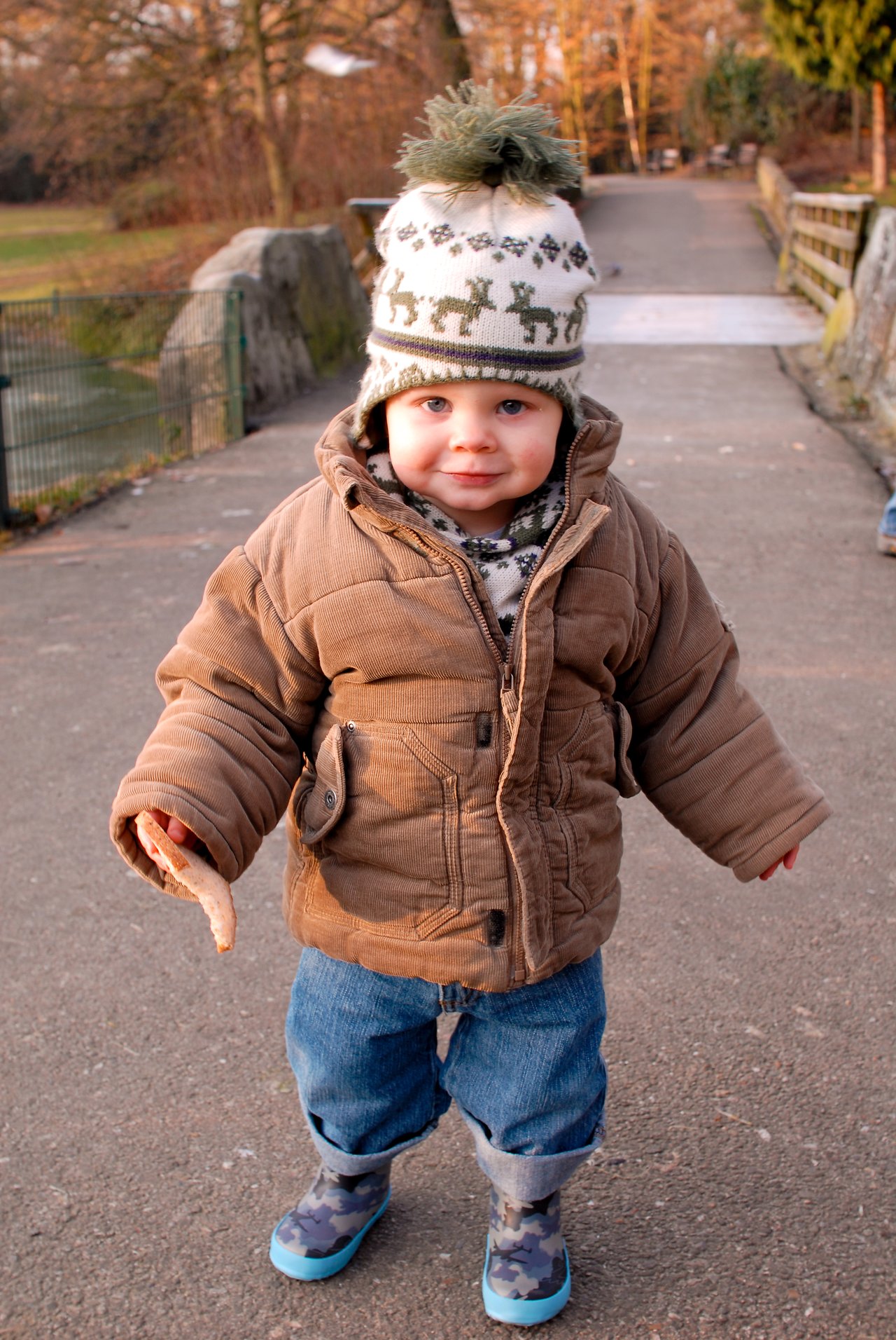 A young child in a winter coat and hat holds a piece of bread, likely for feeding ducks.