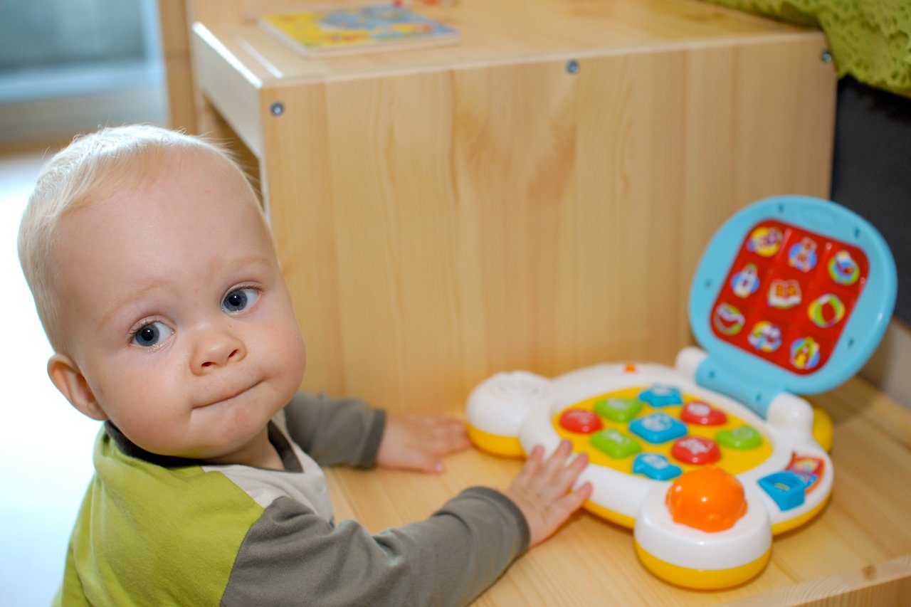 A baby sits at a wooden table, touching a colorful toy laptop with large buttons and a bright screen.