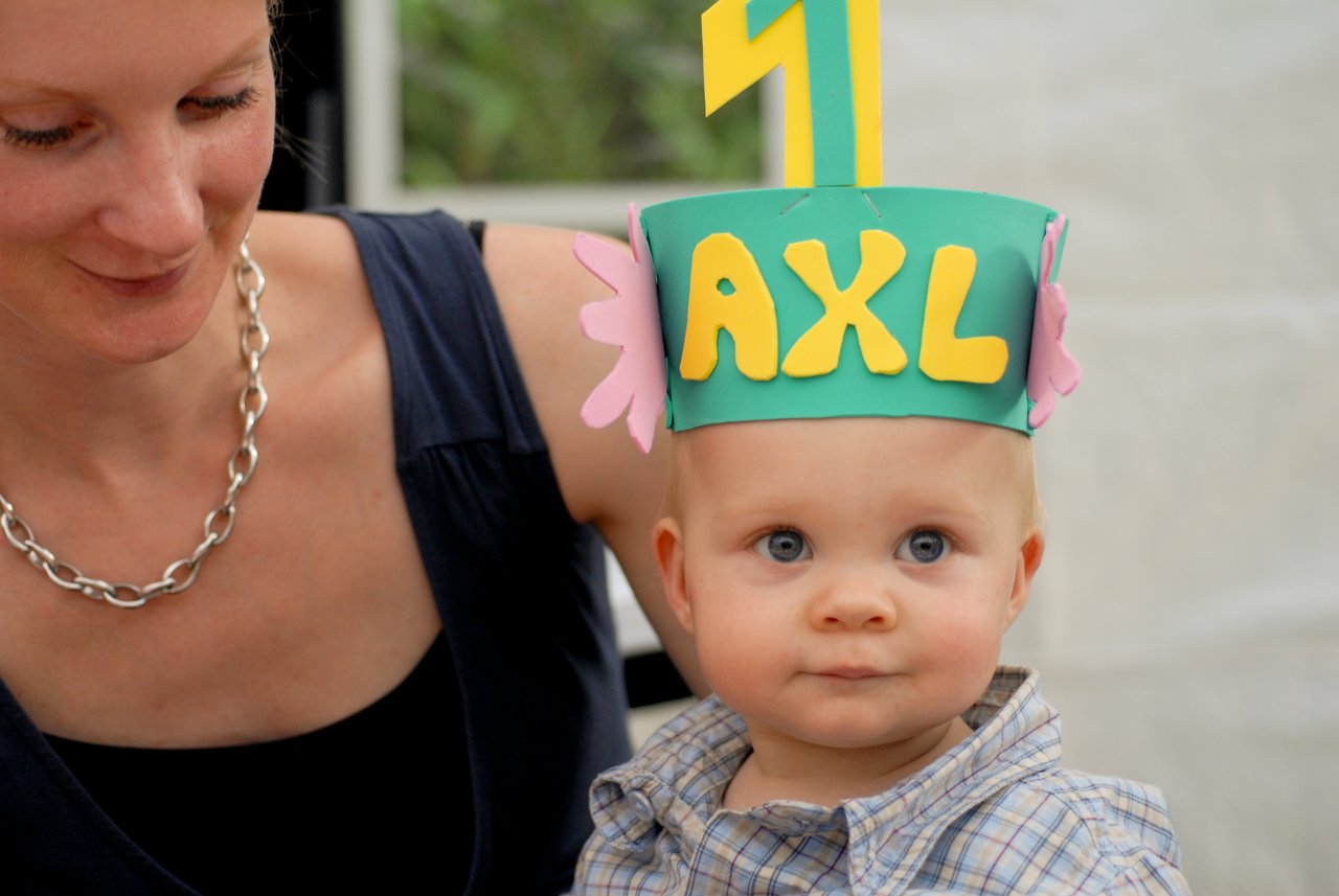A baby wearing a colorful birthday hat with "AXL" and the number one, sitting next to a smiling adult.