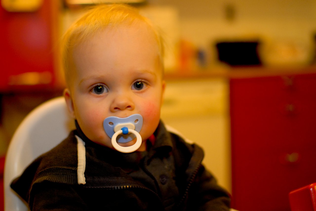 A baby with a blue pacifier sits in a high chair, looking directly at the camera.