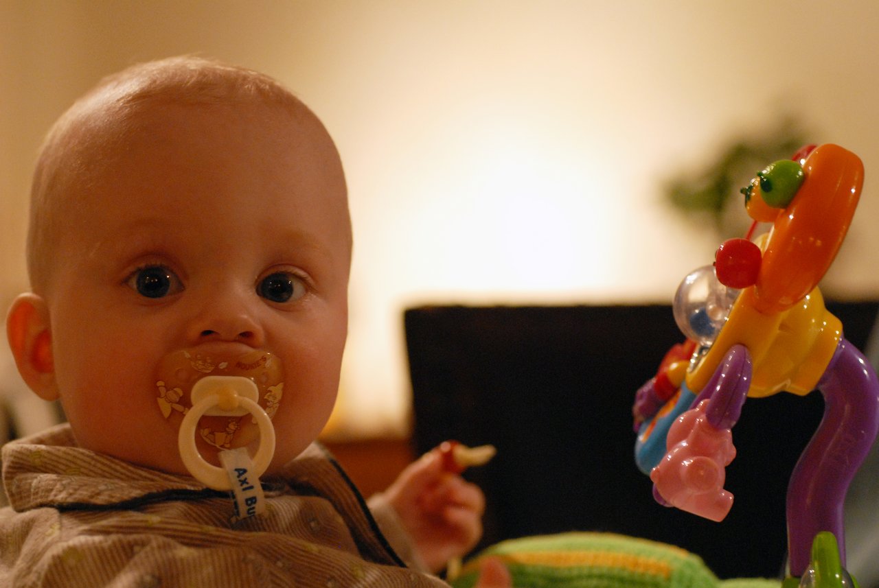 A baby with a pacifier looks toward the camera while holding a snack and playing with a colorful toy.