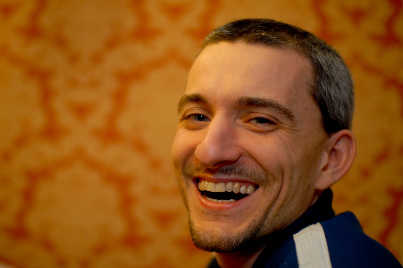 A man with short hair and a beard is smiling broadly, showing his teeth, against a patterned background.