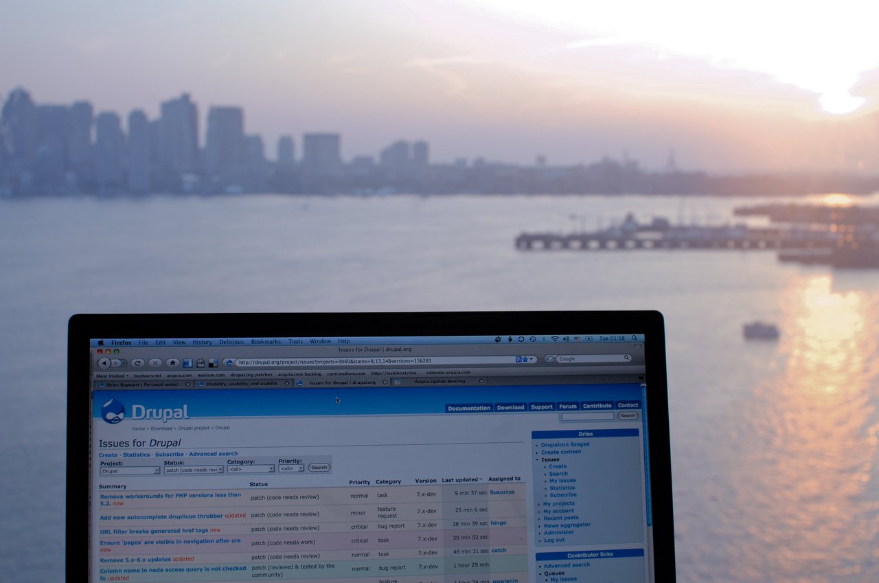 A laptop screen displays a Drupal issue tracker for patch review, with a city skyline and water in the background.