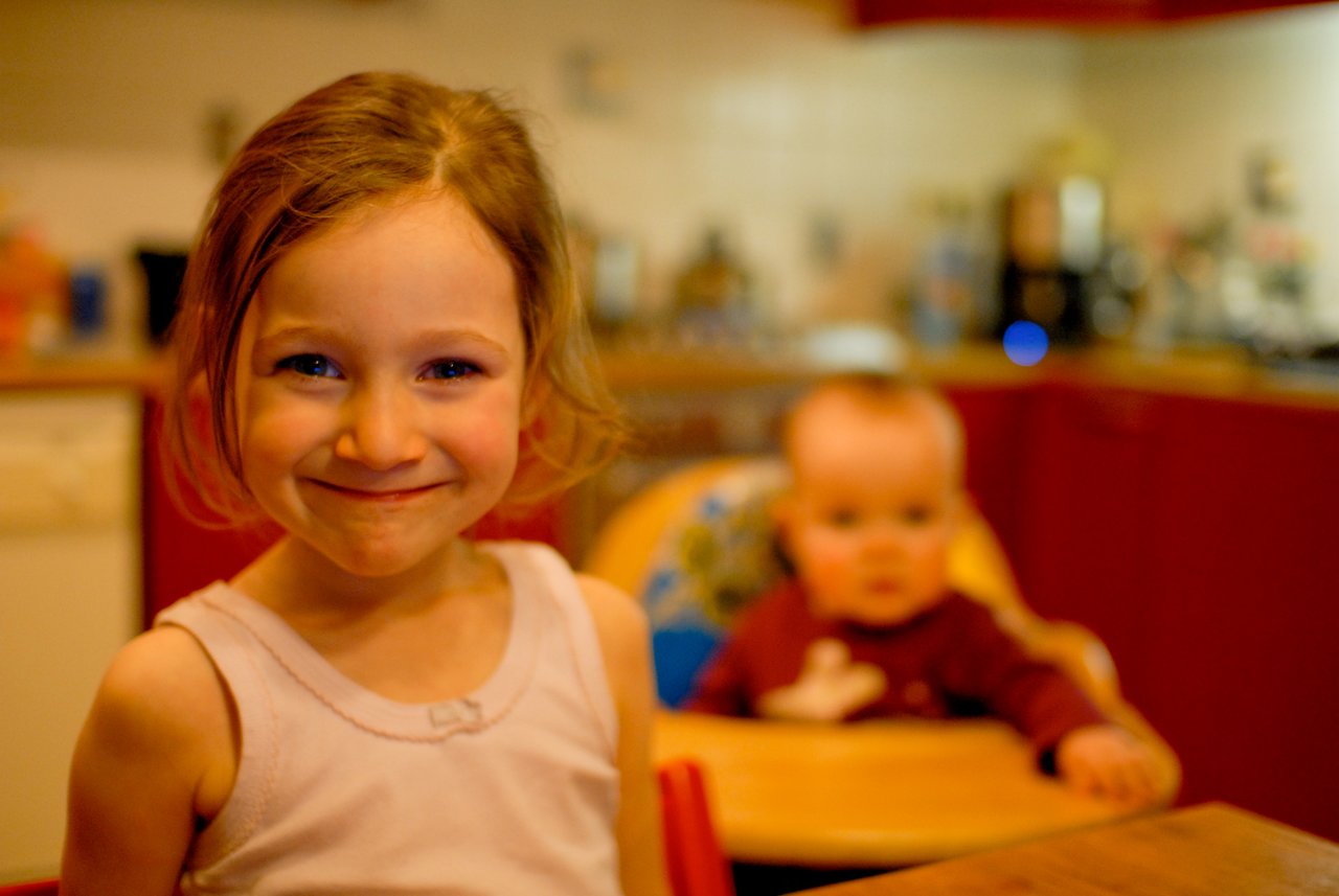 A young girl smiles at the camera while a baby sits in a high chair in the background.
