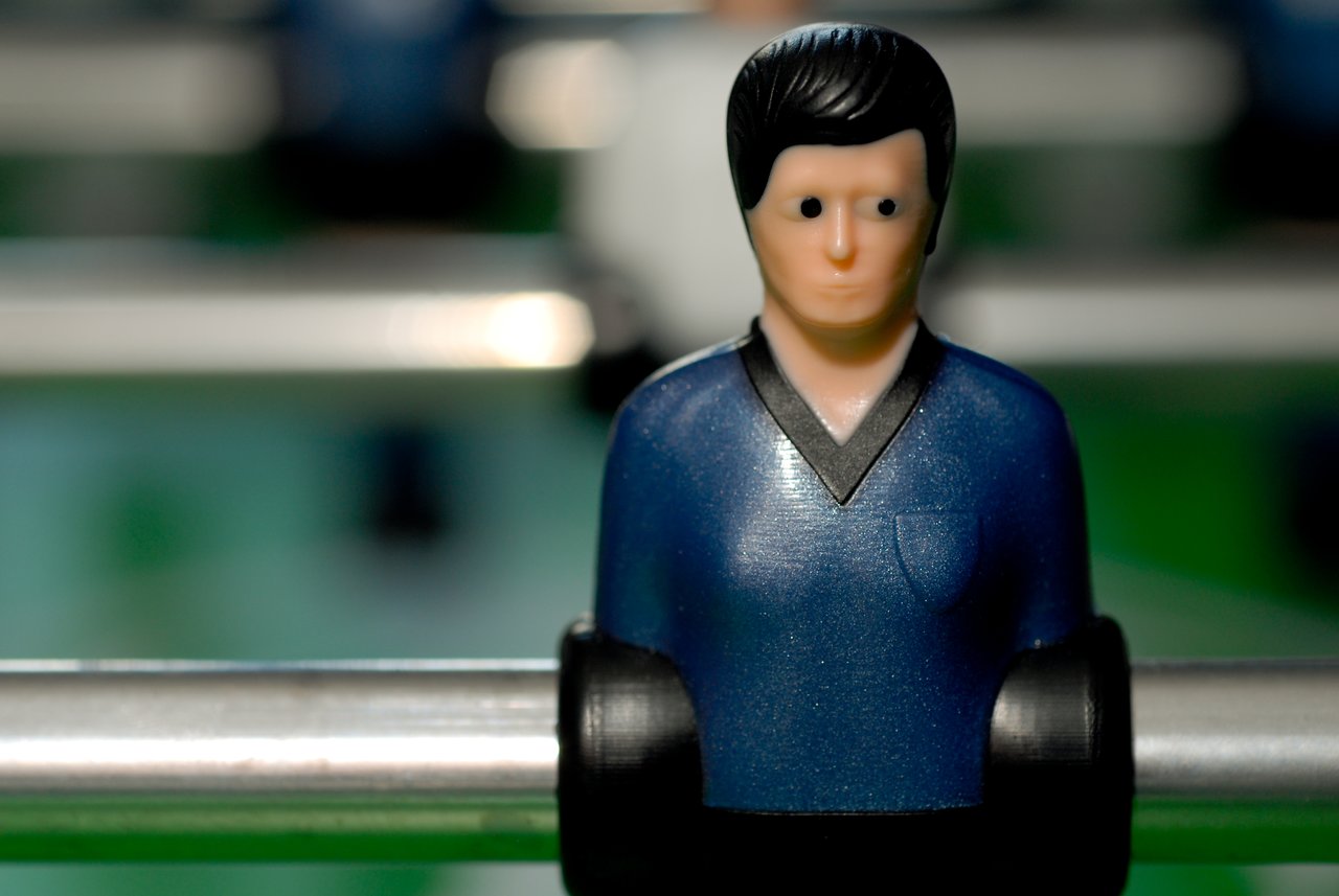 Close-up of a foosball player figure wearing a blue jersey, attached to a metal rod on a green table.