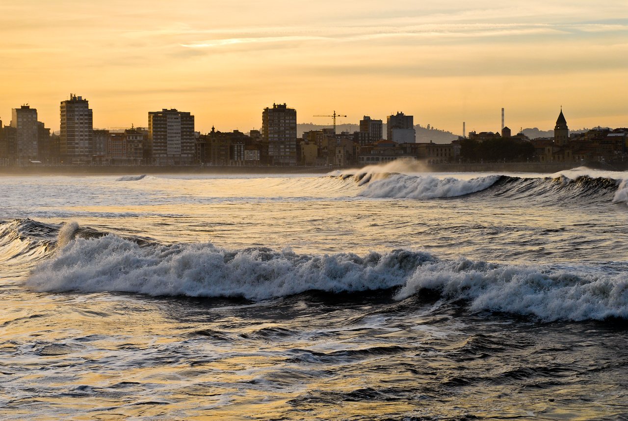 Waves crash against the shore in Gijón, with a city skyline in the background during sunset.