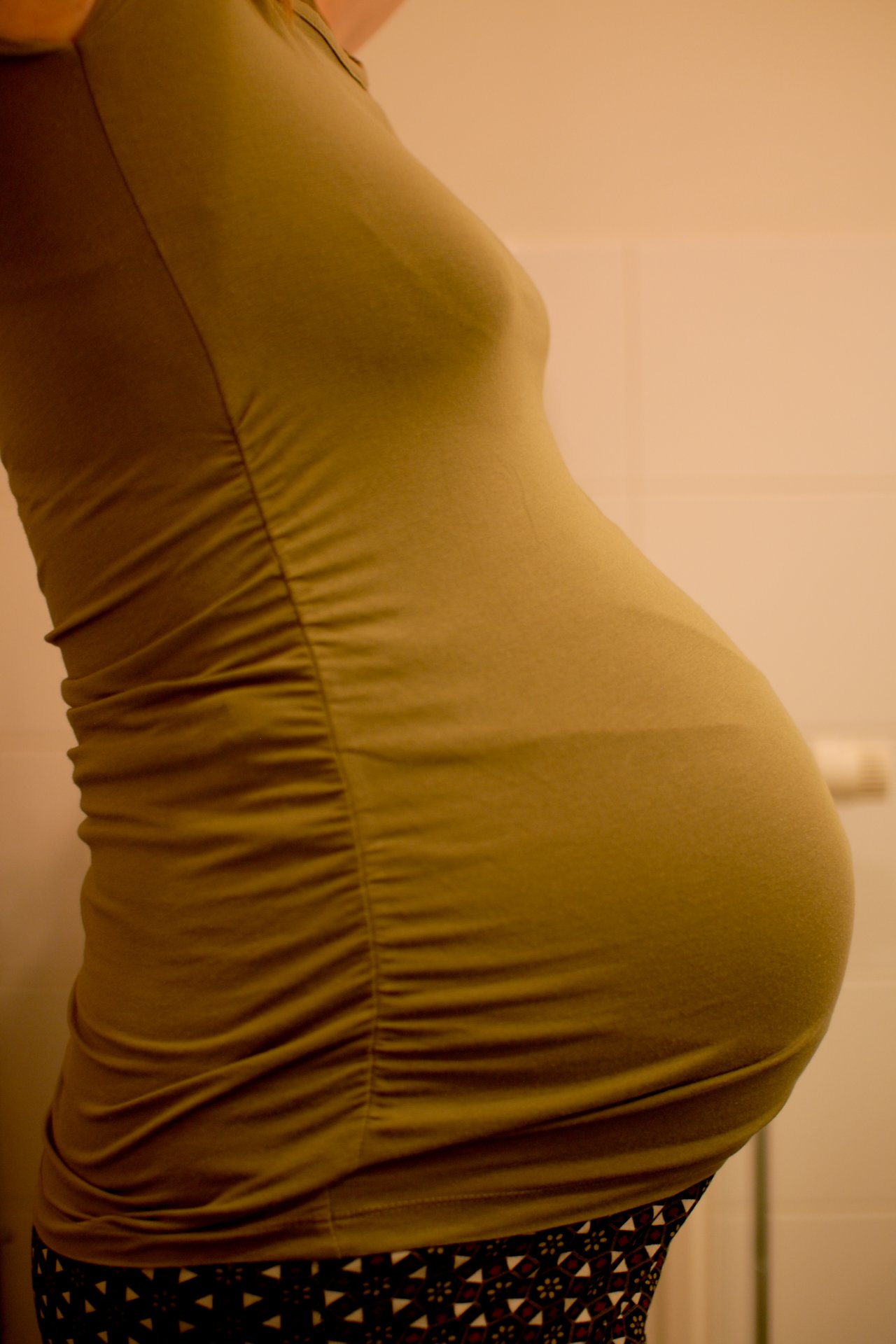 Side view of a pregnant person at 39 weeks, wearing a fitted olive-green top in an indoor setting.