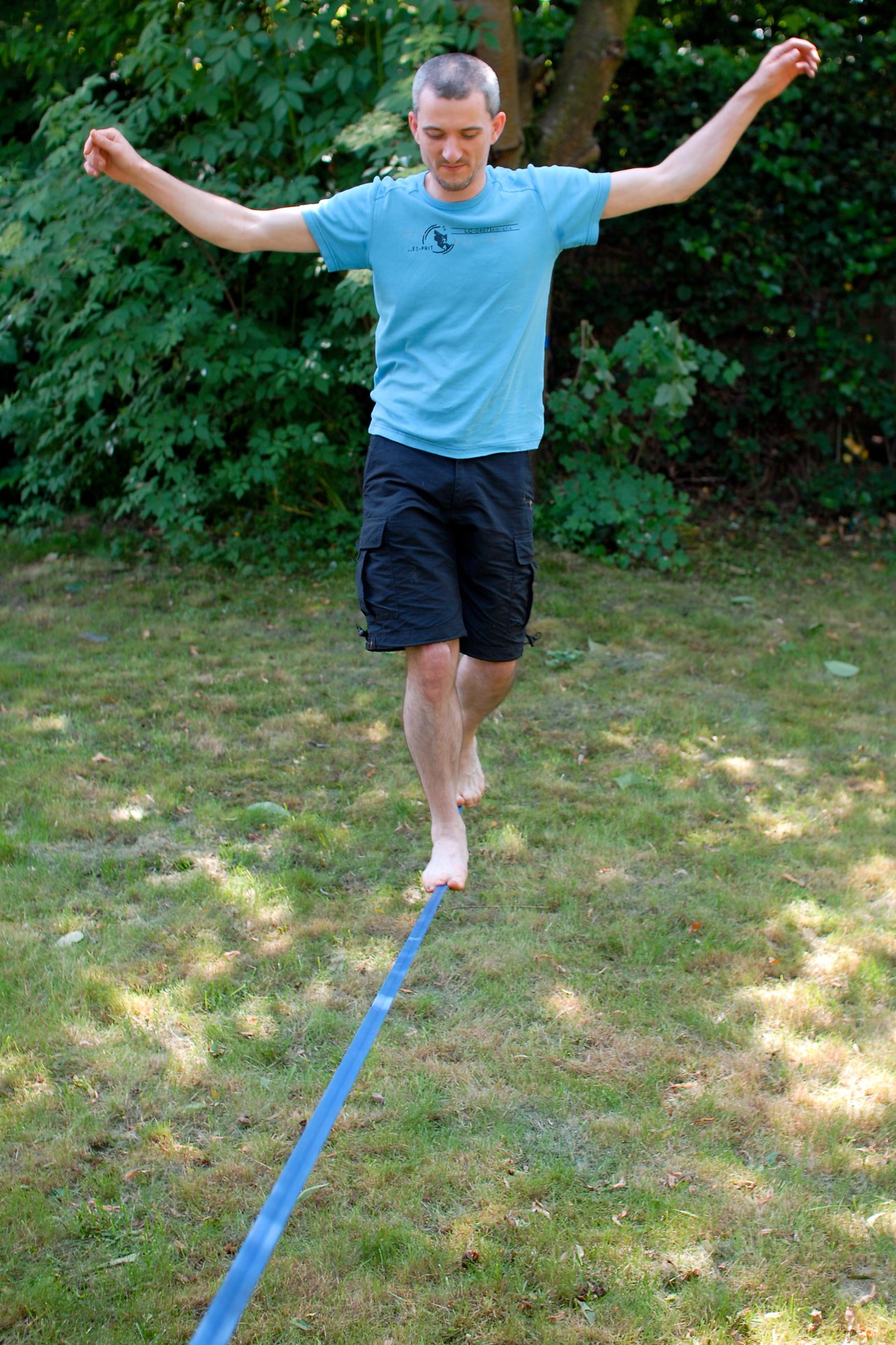 A man in a blue shirt and black shorts balances barefoot on a slackline in a grassy yard.